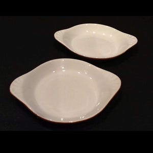 May include: Two white ceramic serving dishes with a brown rim and scalloped edges. The dishes have a shallow, oval shape and are set against a dark background. These dishes are suitable for serving appetizers or side dishes.