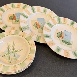 May include: A set of four ceramic plates with a painted leaf and geometric design. The plates have a cream-colored base with green and peach-colored stripes around the edges. The plates are decorated with a leaf and geometric shapes.