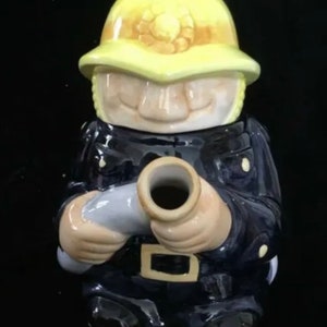 Puede incluir: Un tarro de galletas de cer&aacute;mica con forma de bombero. El tarro representa a un bombero con uniforme azul marino y casco amarillo. El bombero sostiene una manguera gris. El tarro tiene un acabado brillante.