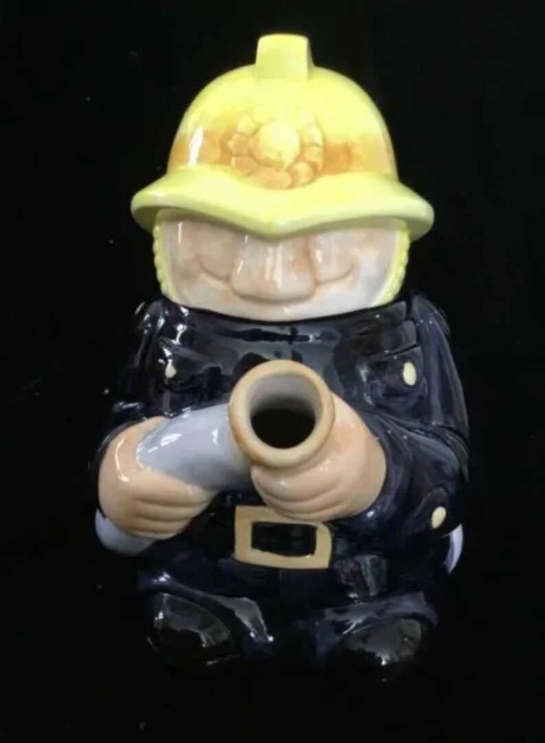 Puede incluir: Un tarro de galletas de cer&aacute;mica con forma de bombero. El tarro representa a un bombero con uniforme azul marino y casco amarillo. El bombero sostiene una manguera gris. El tarro tiene un acabado brillante.