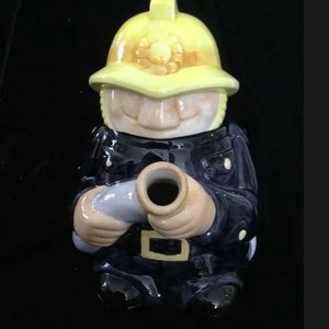 Puede incluir: Figura de cer&aacute;mica de un bombero con uniforme azul marino y casco amarillo. La figura sostiene una manguera gris y tiene una cara sonriente. La figura es un objeto decorativo.