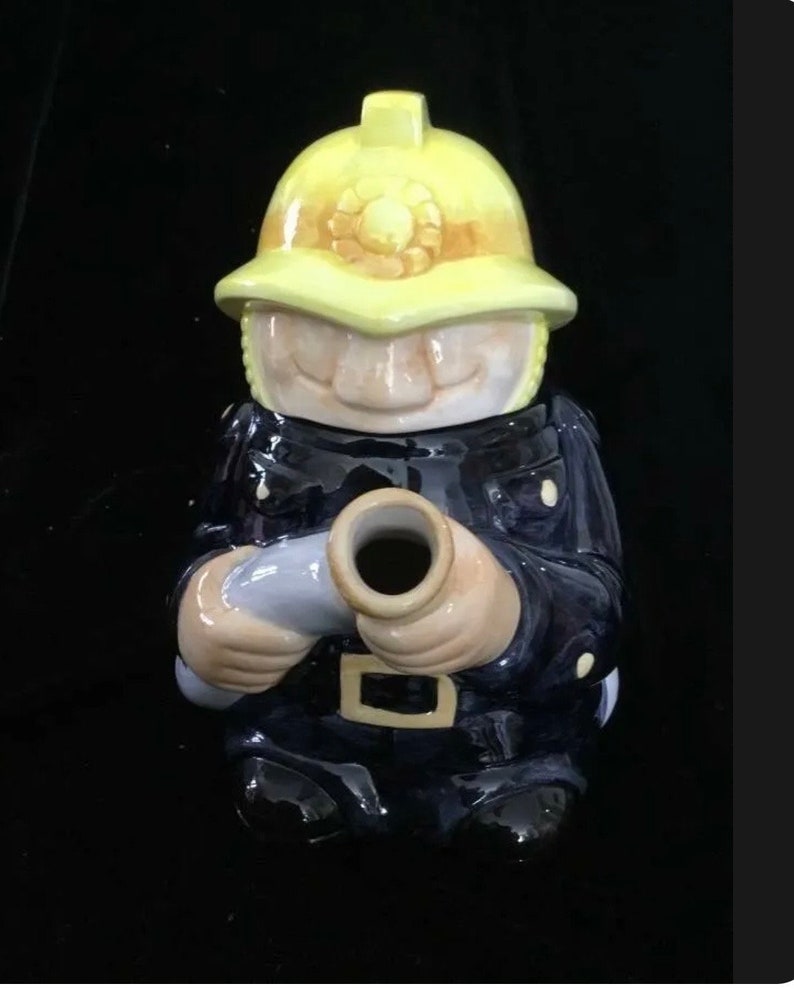 Puede incluir: Figura de cer&aacute;mica de un bombero con uniforme azul marino y casco amarillo. La figura sostiene una manguera gris y tiene una cara sonriente. La figura es un objeto decorativo.