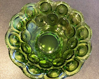 May include: A green glass bowl with a scalloped edge and a starburst pattern. The bowl has a deep green color and a circular shape with a central indentation. The bowl is likely vintage and suitable for serving or display.