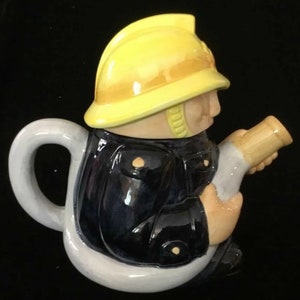 Puede incluir: Una tetera novedosa con forma de bombero. La tetera est&aacute; pintada con un casco amarillo, una chaqueta azul marino y tonos de piel beige. El asa y la base son blancas. El bombero sostiene una manguera beige.
