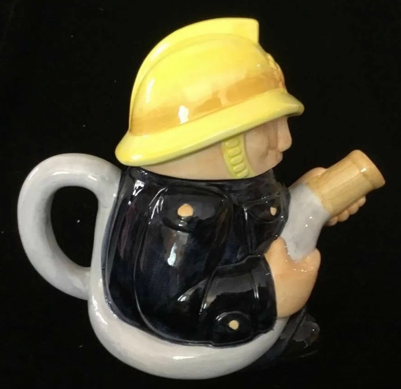 Puede incluir: Una tetera novedosa con forma de bombero. La tetera est&aacute; pintada con un casco amarillo, una chaqueta azul marino y tonos de piel beige. El asa y la base son blancas. El bombero sostiene una manguera beige.
