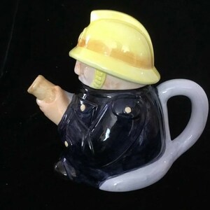 Puede incluir: Tetera de cer&aacute;mica con forma de bombero. La tetera presenta un casco amarillo, una chaqueta azul marino y un asa de color claro. El pico est&aacute; dise&ntilde;ado para parecer un rollo de papel.