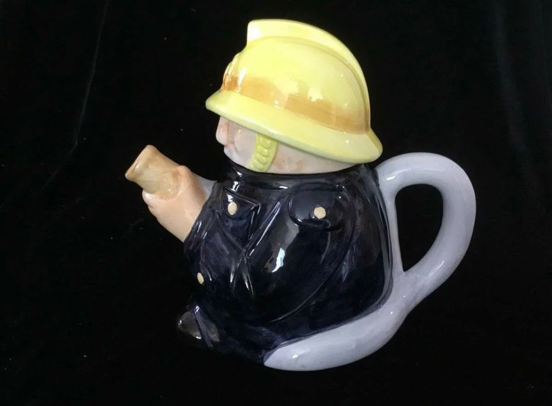 Puede incluir: Tetera de cer&aacute;mica con forma de bombero. La tetera presenta un casco amarillo, una chaqueta azul marino y un asa de color claro. El pico est&aacute; dise&ntilde;ado para parecer un rollo de papel.
