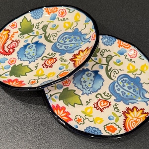 May include: Two white ceramic plates with a blue, yellow, orange, and green floral pattern. The plates have a dark blue rim.