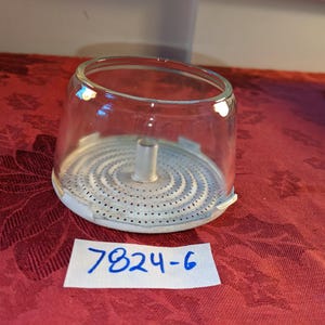 May include: A clear glass dome-shaped cover with a white metal base featuring a perforated circular pattern and a central cylindrical post. A white rectangular paper tag with the handwritten blue ink text 7824-6 is placed in front.