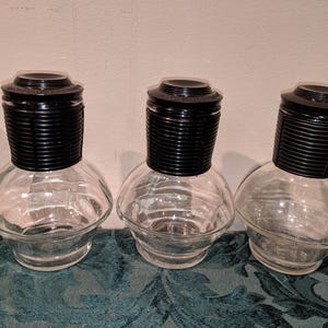 Könnte beinhalten: Drei klare Glasflaschen mit schwarzen gerippten Deckeln. Die Flaschen sind leer und haben eine runde Form.