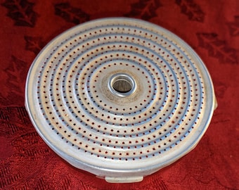 Op de afbeelding: Een cirkelvormig metalen douchescherm voor een espressomachine met een fijn patroon van kleine gaatjes in concentrische cirkels, een centrale opening en zichtbare sporen van lichtbruine resten.