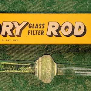 Pode incluir: Uma vareta de filtro de vidro transparente com uma ponta afiada e uma seção central bulbosa, posicionada abaixo de uma caixa retangular amarela com texto preto que diz CORY GLASS FILTER ROD e REG. U. S. PAT. OFF. A vareta é lisa.