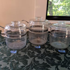 Puede incluir: Tres cafeteras de vidrio transparente con tapas y asas. Cada olla tiene una banda plateada en el medio. Las ollas están sobre un mantel verde y negro estampado. Pequeñas etiquetas azules están adheridas a la parte inferior de las ollas.