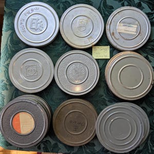May include: A collection of vintage film canisters in various shades of gray and silver. Some canisters have labels or text, including "South Pacific" and "OA 4D". The canisters are arranged on a patterned green surface.