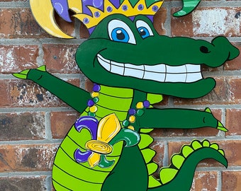 May include: A wooden alligator decoration with a Mardi Gras theme. The alligator is painted green with a yellow crown, purple and yellow hat, and a necklace of beads. It has a large smile and outstretched arms.