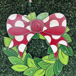 May include: A red and white polka dot bow with a green wreath. The bow is tied with a red ribbon and the wreath is made of green leaves.