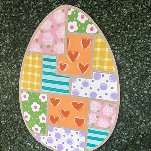 May include: A wooden Easter egg-shaped wall hanging with a patchwork design. The design includes squares of pink roses, green flowers, orange hearts, yellow plaid, blue stripes, and purple polka dots. A wire and wooden beads are attached for hanging.