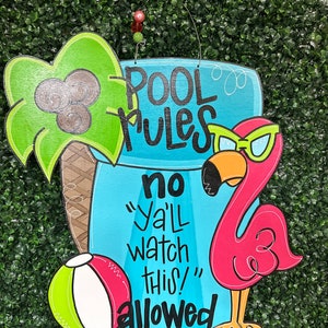 May include: A wooden sign with a blue background and the text "Pool Rules" in black. The sign also has a pink flamingo with sunglasses, a green palm tree, and a beach ball. The text "no "ya'll watch this!" allowed" is written in black.