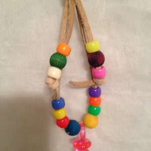 May include: A colorful beaded necklace with a pink pacifier charm. The necklace is made of brown leather cord and features a variety of colored beads, including red, orange, yellow, green, blue, and purple.
