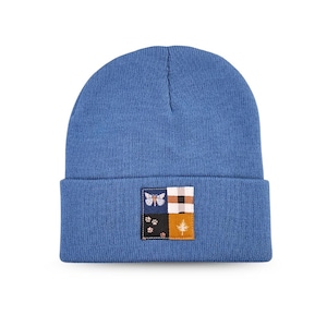 May include: A blue knitted beanie hat with a square patch. The patch features a butterfly, a plaid pattern, paw prints, and a tree. The hat is a solid blue colour and has a folded brim.