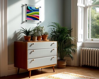 May include: A wooden dresser with three light gray drawers and small round knobs, topped with four potted plants. Above the dresser hangs a square abstract painting in a clear acrylic frame featuring vibrant wavy lines of blue, green, and pink.