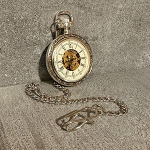 Può includere: Un orologio da tasca color argento con quadrante scheletrato che mostra ingranaggi interni dorati e numeri romani neri. L'orologio presenta un bordo esterno testurizzato ed è collegato a una catena in metallo argentato con chiusura.
