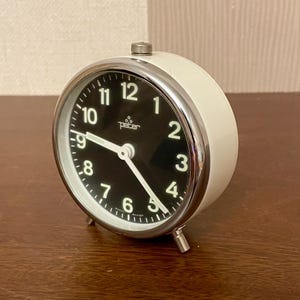 Könnte beinhalten: Ein weiß-silberner Wecker im Vintage-Stil mit schwarzem Zifferblatt. Die Uhr hat eine silberne Lünette und Beine, mit dem Wort "peter" auf dem Zifferblatt. Die Uhr steht auf einer Holzoberfläche.