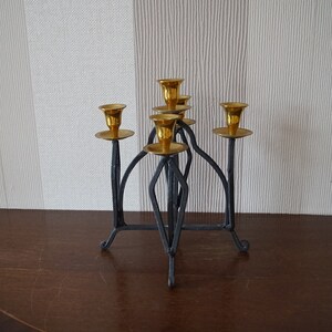 May include: A black metal candelabra with five gold candle holders. The candelabra has a geometric design and stands on a wooden surface.