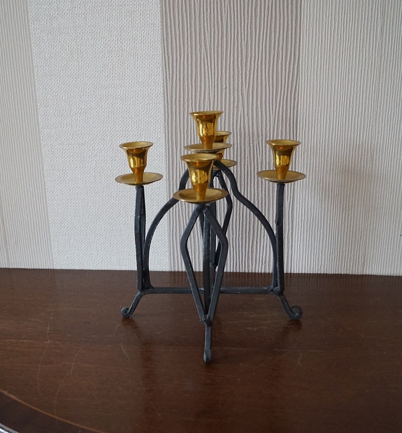 May include: A black metal candelabra with five gold candle holders. The candelabra has a geometric design and stands on a wooden surface.