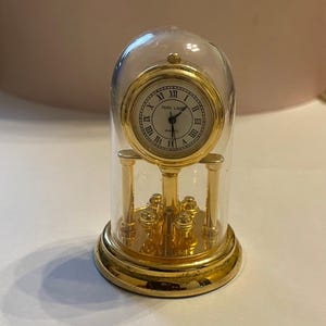 Op de afbeelding: Een kleine, goudkleurige quartz klok met een witte wijzerplaat en zwarte Romeinse cijfers, ingekapseld in een heldere glazen koepel. De klok heeft een gouden basis en steunpilaren. De wijzerplaat leest "PARK LANE QUARTZ".
