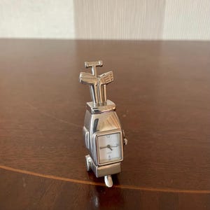 Peut inclure: Une montre à quartz argentée en forme de sac de golf avec des clubs. Le cadran de la montre est carré avec des chiffres et des aiguilles noirs. La montre mesure environ 5 cm de haut.