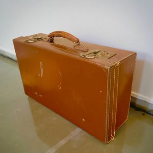 Pode incluir: Uma mala castanha vintage com uma pega de couro e fechos cor de latão. A mala apresenta sinais de desgaste, com bordas costuradas e formato retangular. A estética geral sugere um acessório de viagem clássico.