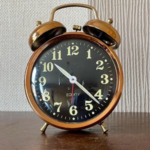 May include: A round analog alarm clock with a metallic copper-colored frame and two bells on top. The clock face is black with white Arabic numerals from 1 to 12, white hour and minute hands, a red second hand, and the word EQUITY printed in white.