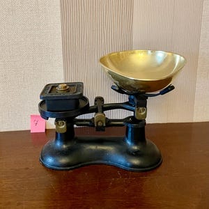 Könnte beinhalten: Eine antike schwarz-goldene Waage mit einer Messingschale und einer quadratischen Plattform. Die Waage hat ein klassisches Design mit einem gebogenen Sockel und einer Reihe von Hebeln und Gewichten. Ein kleiner rosa Haftzettel mit der Zahl 7 ist angebracht.