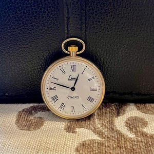 Op de afbeelding: Een goudkleurig Limit International quartz zakhorloge met een witte wijzerplaat en zwarte Romeinse cijfers. Het horloge heeft een gouden opwindstang en een kleine lus voor een ketting. Het horloge staat tegen een zwarte lederen achtergrond.