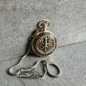 Può includere: Un orologio da tasca color argento con un emblema a forma di aquila in rilievo sul coperchio anteriore. L'orologio è fissato a una catena flessibile a serpente che include una clip in metallo all'estremità per il fissaggio agli abiti.