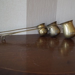 May include: Three antique brass measuring cups with long handles. The cups are graduated in size and have a rounded shape. The handles are attached to the cups with a loop.