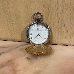 Puede incluir: Un reloj de bolsillo plateado con esfera blanca, números y manecillas negras. La esfera muestra el texto "SWISS BALANCE" y la cruz suiza. La tapa dorada está abierta, revelando el interior texturizado.