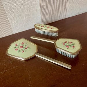 Könnte beinhalten: Ein Toilettenset bestehend aus einem Handspiegel, einer Haarbürste und einer Kleiderbürste. Jedes Teil hat eine hellgrüne Emaille-Rückseite mit einem Blumenmuster aus rosa Rosen und grünen Blättern, eingefasst in geriffeltem, goldfarbenem Metall.
