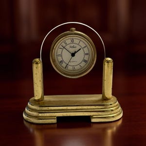 Puede incluir: Un reloj de escritorio de cuarzo dorado con esfera blanca y números romanos negros. El reloj tiene un arco de cristal curvado y una base rectangular. La esfera del reloj dice "Reflex" y "Quartz".