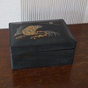 May include: A black lacquered wooden box with a gold and silver design of a tiger in a forest setting. The box has a hinged lid and is decorated with a moon and clouds.