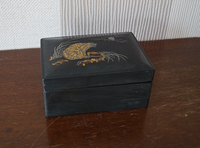 May include: A black lacquered wooden box with a gold and silver design of a tiger in a forest setting. The box has a hinged lid and is decorated with a moon and clouds.