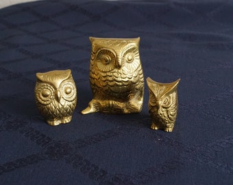 May include: Three gold-colored owl figurines. The largest owl is sitting with its wings folded. The two smaller owls are standing upright. The owls are all facing forward.