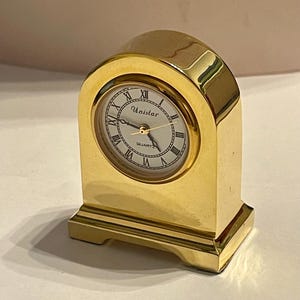 Puede incluir: Un reloj de cuarzo Unistar de color dorado con base rectangular y parte superior arqueada. La esfera es blanca con números romanos negros y agujas doradas. El reloj está hecho de un material brillante y reflectante.