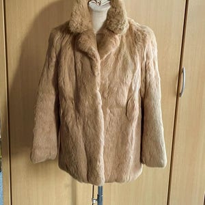 Puede incluir: Un abrigo de piel sintética de color beige con un cuello ancho, exhibido en un perchero de metal. El abrigo tiene un aspecto suave y texturizado y un diseño clásico. El abrigo se exhibe frente a un armario de madera marrón claro.