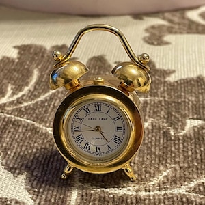Può includere: Una piccola sveglia al quarzo dorata con quadrante bianco e numeri romani neri. L'orologio ha una maniglia curva e due campanelli. Sul quadrante si legge "PARK LANE QUARTZ".