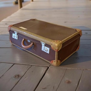 Puede incluir: Una maleta marrón vintage con ribetes y protectores de esquina color beige. La maleta tiene un asa y dos cierres metálicos. La maleta rectangular descansa sobre una superficie de madera.