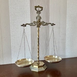Puede incluir: Una balanza de latón con una columna central de textura similar al bambú, una barra transversal ornamentada con volutas decorativas y dos platos circulares suspendidos por finas cadenas de metal en cada extremo del brazo horizontal.