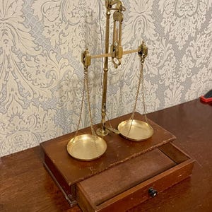 Puede incluir: Una antigua balanza de latón con dos platillos circulares suspendidos por cadenas. La balanza está montada sobre una base de madera con un cajón. Es un artículo vintage, probablemente utilizado para medir pequeñas cantidades.