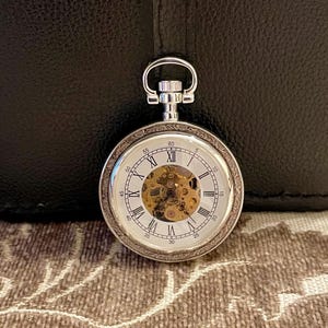 Op de afbeelding: Zilveren zakhorloge met een witte wijzerplaat, Romeinse cijfers en een zichtbaar mechanisch uurwerk. Het horloge heeft een decoratieve rand en een lus voor een ketting. De achtergrond is een textuur.